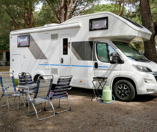 A white RV parked under pines, with camp chairs and a small table set out beside it.