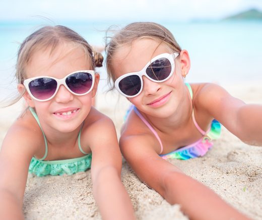 Two girls in sunglasses lie on the sand, smiling at the camera.