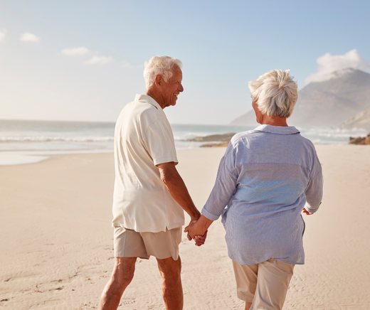 An older couple walks hand in hand along a sunny beach.