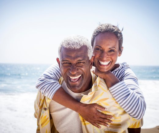 A smiling couple poses on the beach, one with an arm around the other’s shoulders.