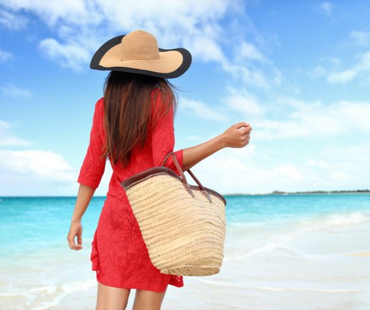 A woman in a red dress and a wide straw hat walks the shoreline with a woven beach bag.